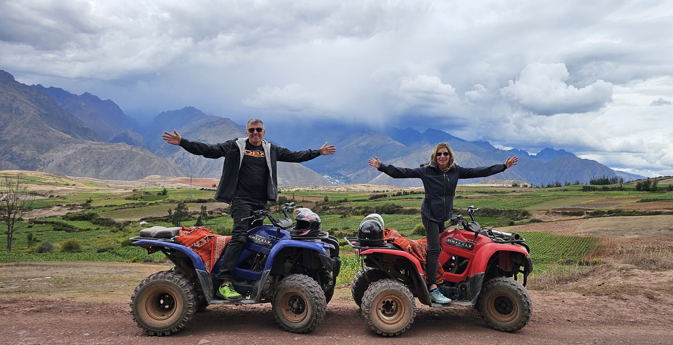 Chip and his wife standing arms-wide on two ATVs on a dirt road in the Peruvian Andes, storm clouds and green valley behind them